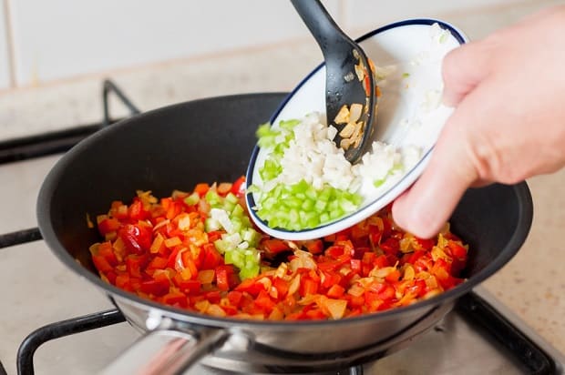 зеленый перец с чесноком пересыпается в сковороду с красным перцем и луком на плите