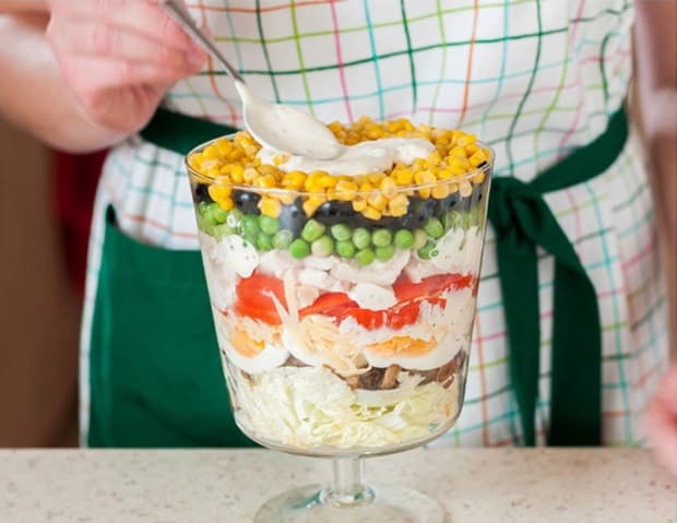 слоеный салат из кукурузы, горошка, яиц, помидоров, маслин и курицы в салатнице на ножке на столе