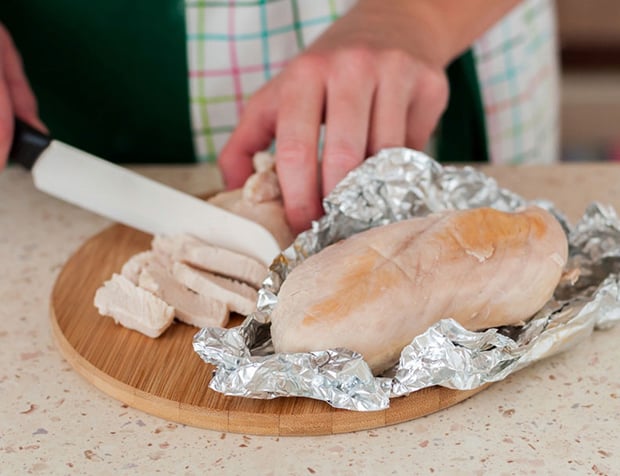 запеченное куриное филе на фольге на круглой разделочной доске на столе, рядом филе нарезается ножом