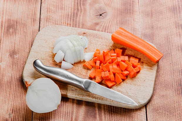 нарезанные морковка и луковица с ножом на разделочной доске, рядом очищенная луковица и морковка