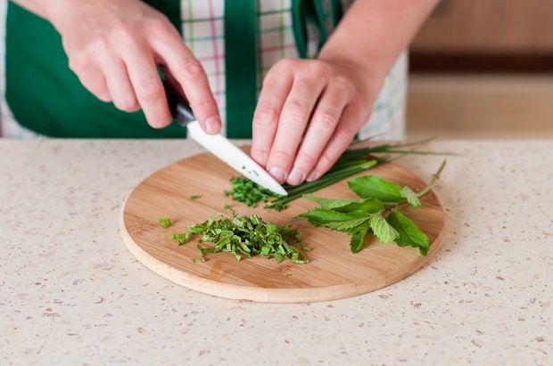 зеленый лук нарезается ножом, рядом веточка мяты на разделочной доске на столе