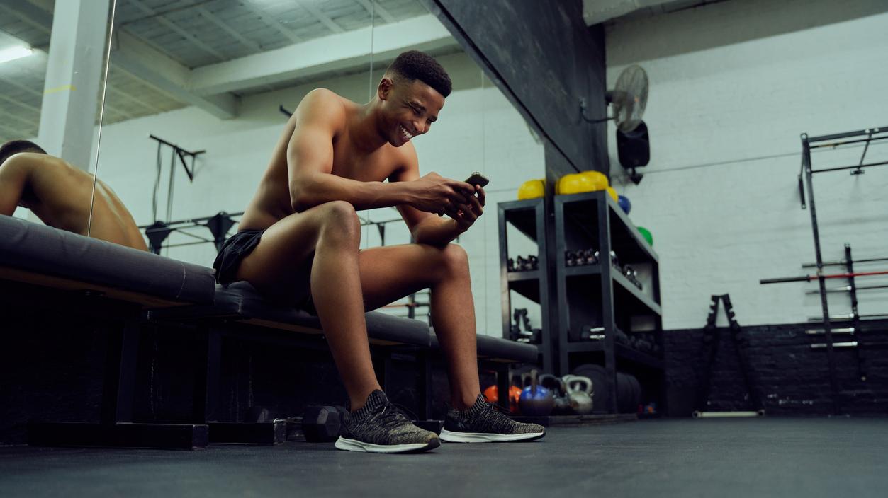 Young African American male looking at his phone to reply to an online message. Happy, mixed race male smiling at his phone while sitting down in the gym.